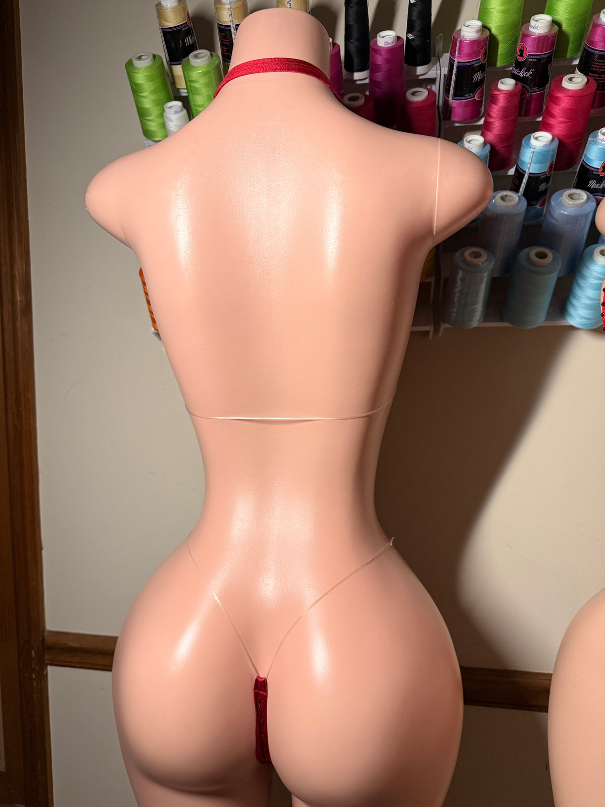 Rear view of a flesh-toned mannequin torso wearing a very minimal red thong-style underwear bottom, standing indoors in front of shelves of colorful thread spools.